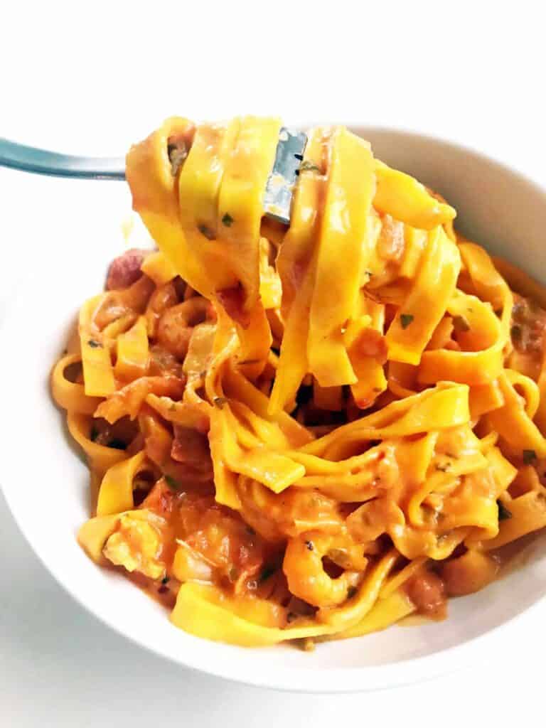 Creamy saffron arrabiata shrimp with tagliatelle with a fork in a white bowl on a white table surface