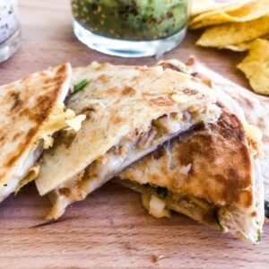 Close up of chicken quesadilla cut in pieces on a wooden choppingboard with guacamole, ranch dip and tortilla chips