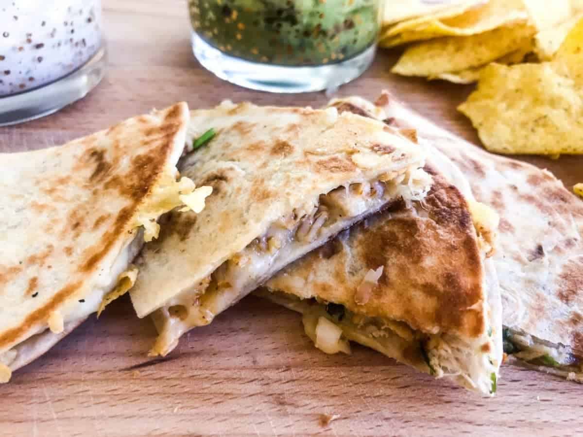 Close up of chicken quesadillas cut in pieces on a wooden choppingboard with guacamole, ranch dip and tortilla chips