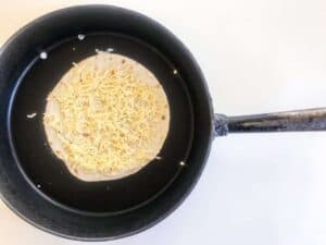 Step two of cooking Chicken Quesadillas - Tortilla topped with cheese in a black pan on a white surface