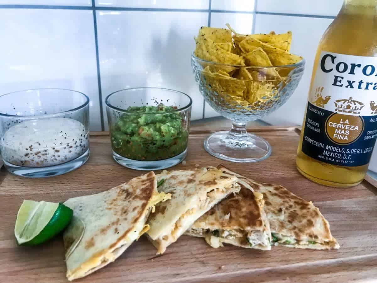 Chicken quesadillas cut in pieces on a wooden chopping board with lime slices, guacamole, ranch dip, tortilla chips and a Corona beer