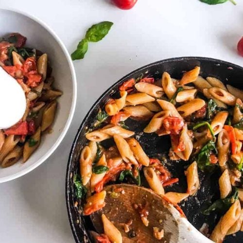 15-minute cherry tomato pasta with spinach and walnuts in a black skillet and in a white bowl topped with burrata, all on a white table decorated with cherry tomatoes on a branch and basil leaves