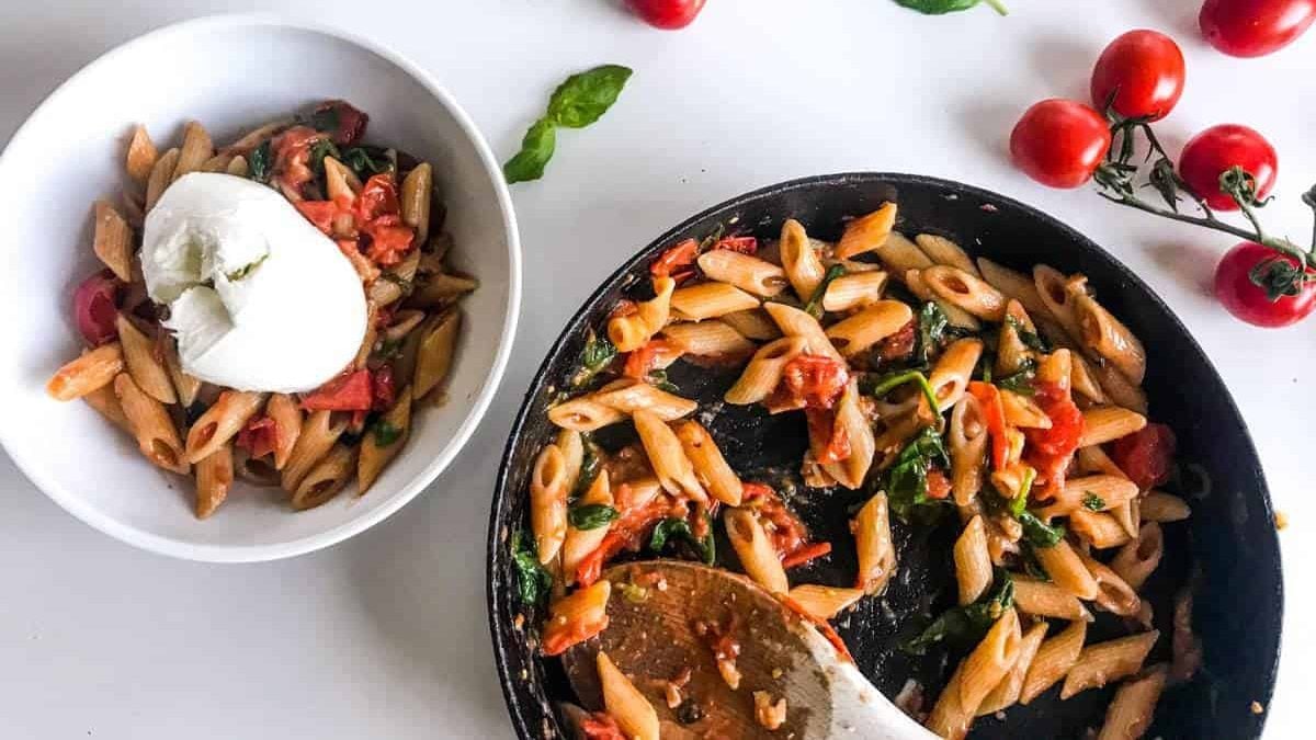 15-minute cherry tomato pasta with spinach and walnuts in a black skillet and in a white bowl topped with burrata, all on a white table decorated with cherry tomatoes on a branch and basil leaves