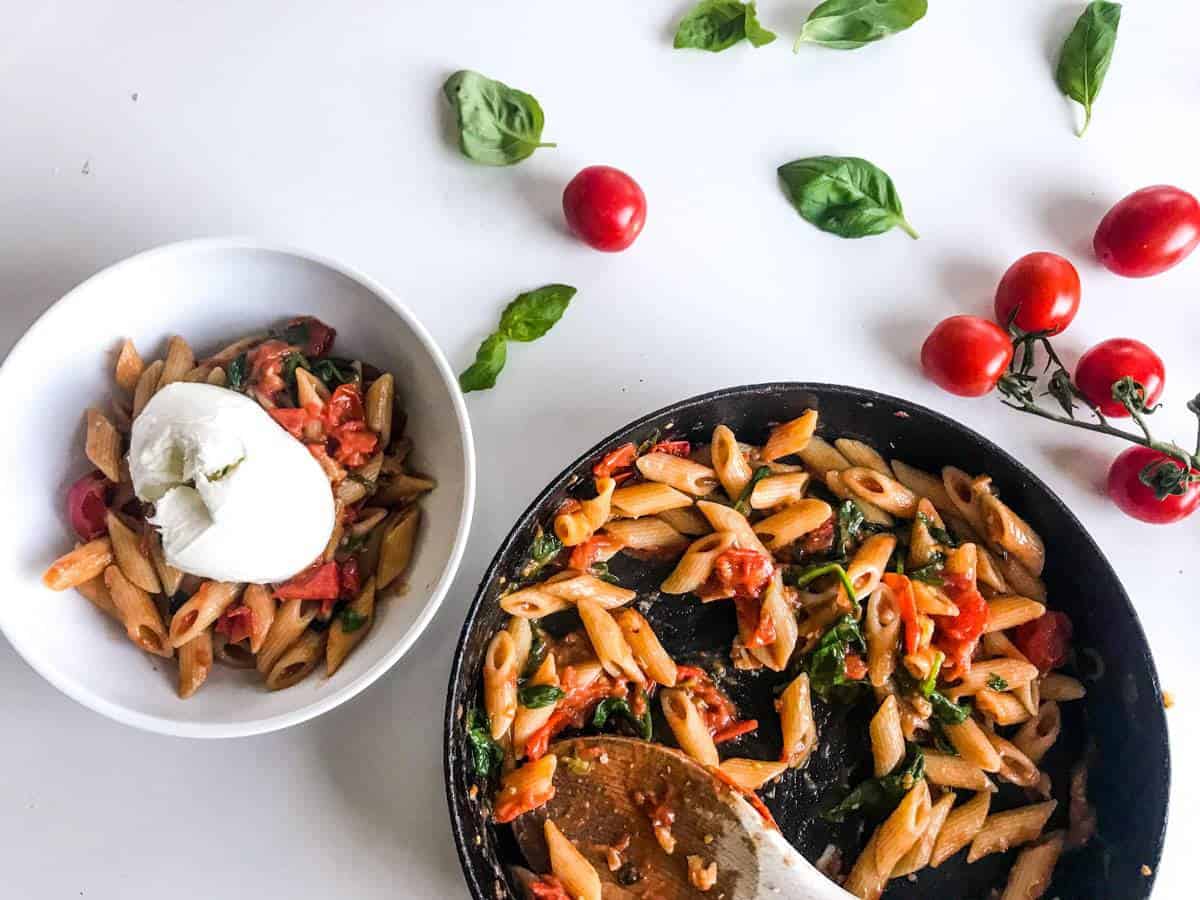 15-minute cherry tomato pasta with spinach and walnuts in a black skillet and in a white bowl topped with burrata, all on a white table decorated with cherry tomatoes on a branch and basil leaves