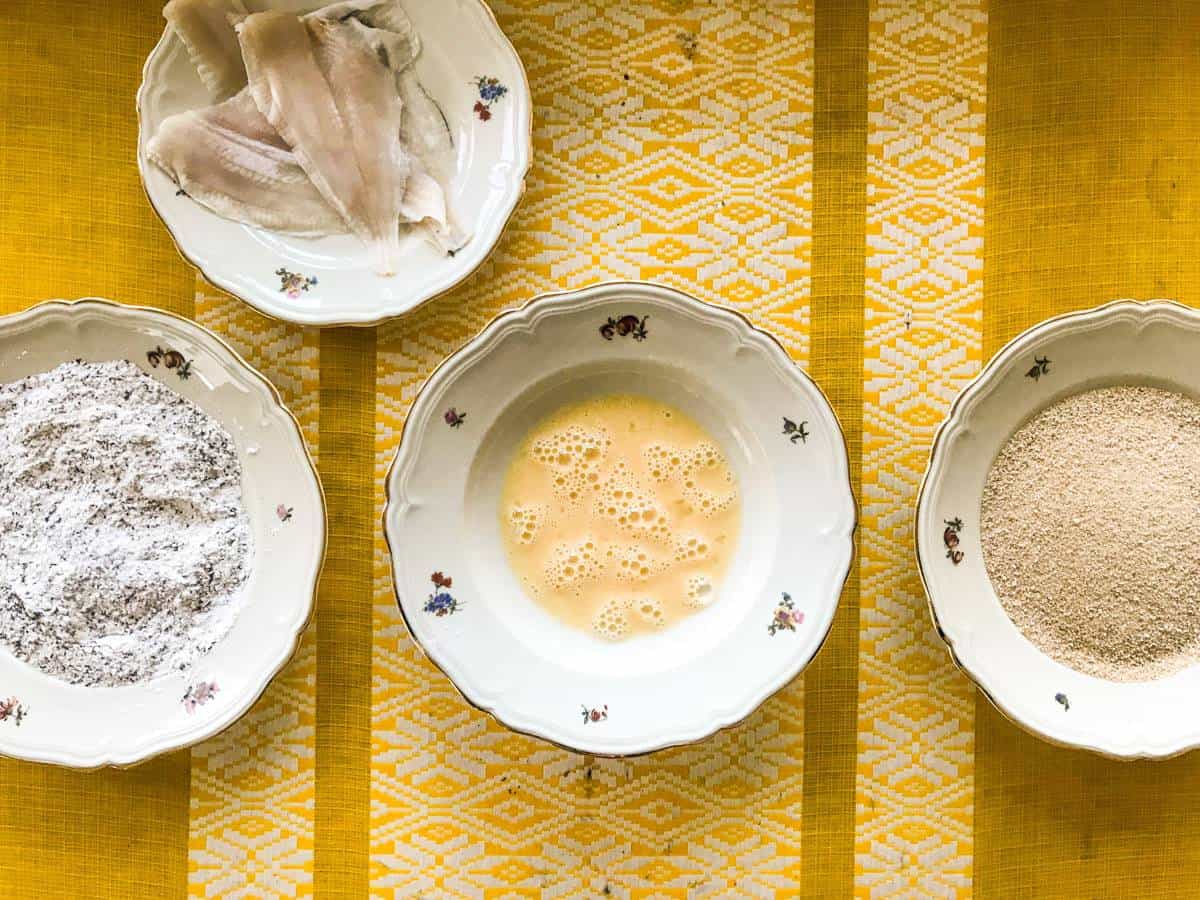 Plaice filets on a plate, basil flour on a plate, whipped egg in a deep plate, breadcrumbs on a plate, all on a yellow table for pan-seared breaded plaice with whipped lemon butter