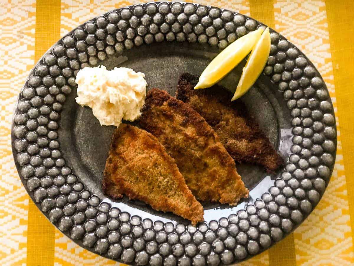 Pan-seared breaded plaice with whipped lemon butter and lemon wedges on a black plate on a yellow table