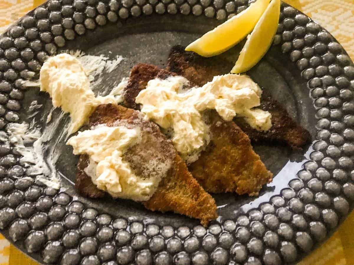 Pan-seared breaded plaice with whipped lemon butter and lemon wedges on a black plate on a yellow table