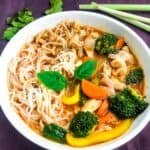 thai chicken noodle soup in a white bowl on a purple surface with lemon grass and kaffir lime leaves