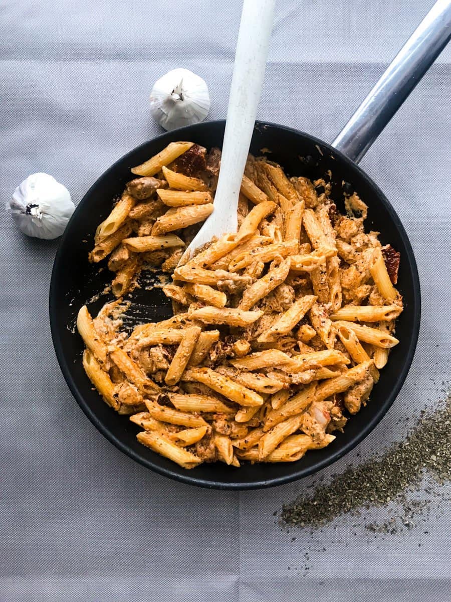 Creamy sun-dried tomato chicken pasta with crème fraiche in a black pan on a grey surfae with two garlics and ground basil
