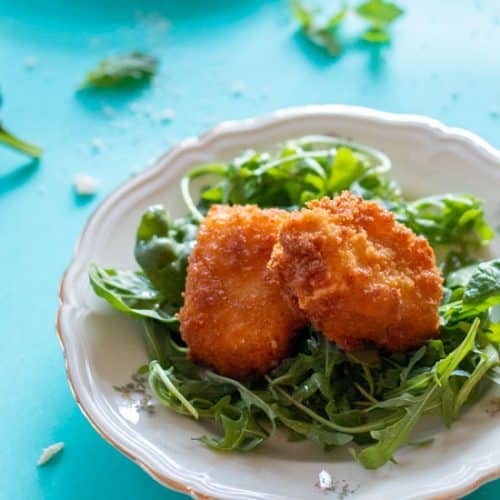 two pieces of panko crusted feta cheese on a bed of greens on a plate, on a turquoise background.