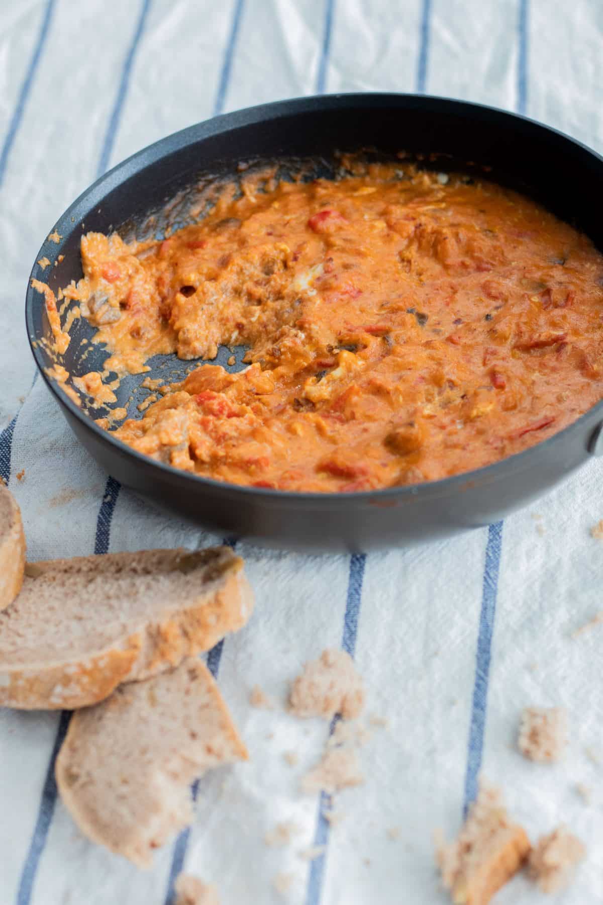 shakshuka in a frying pan on a white and blue striped kitchen towel with a few pieces of bread in front