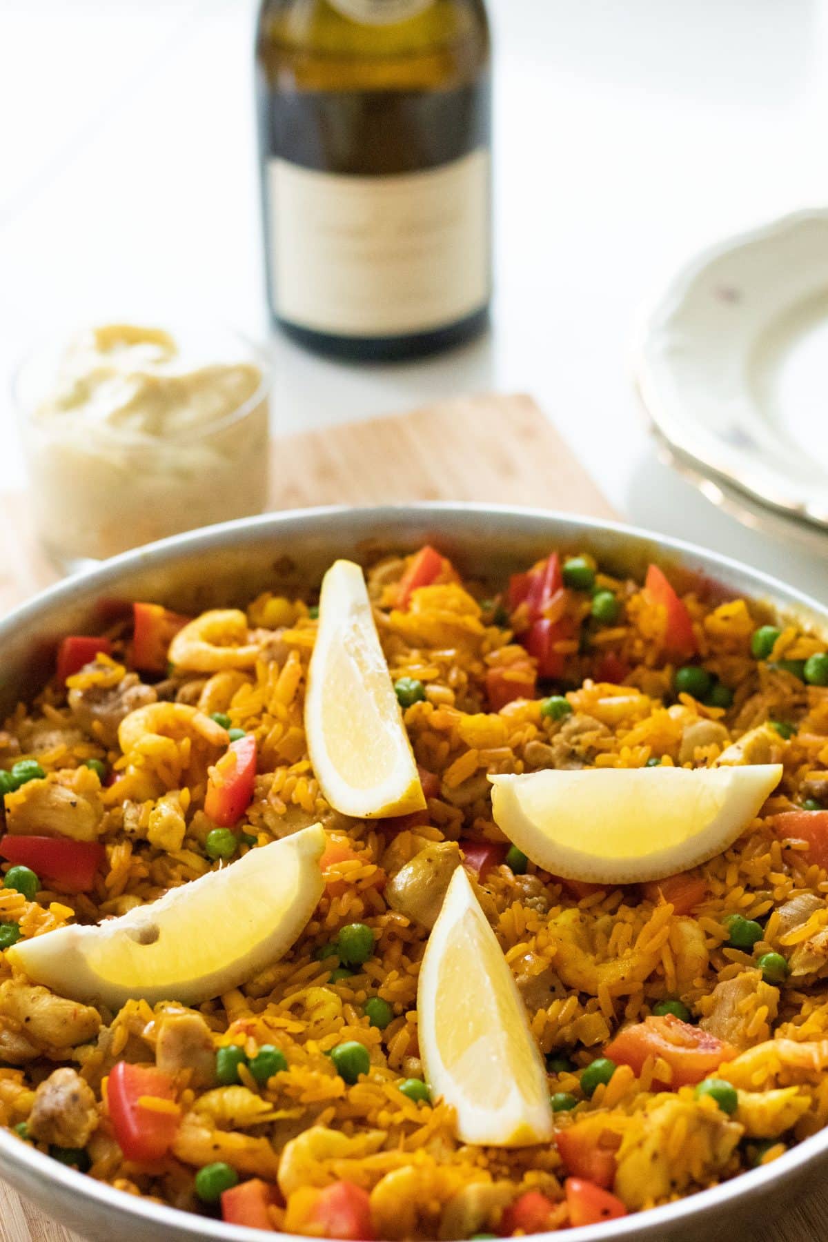 side view of paella with lemon wedges with lime aioli and a bottle of wine in the background