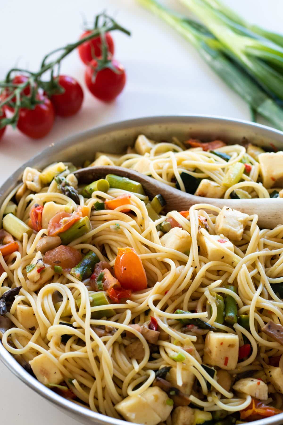 halloumi pasta primavera in a pan with a wooden spoon, with cherry tomatoes and spring onions in the background