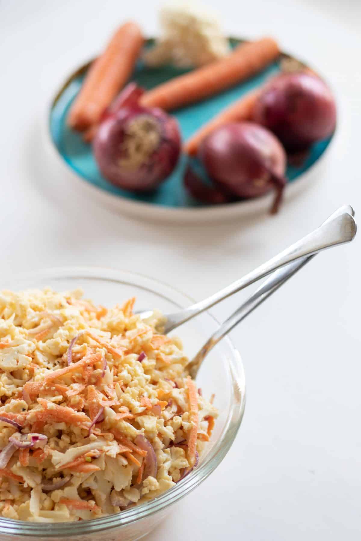 raw cauliflower salad or caulislaw in a glass bowl and a plate with red onions, carrots and cauliflower in the background 