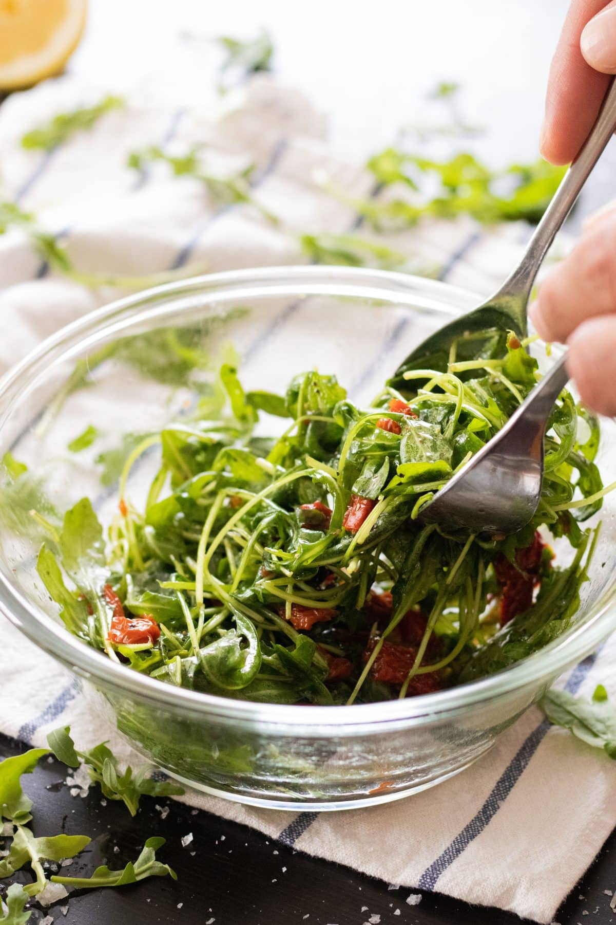 side view of arugula salad with sun-dried tomatoes in a glass bowl with a spoon and fork lifting up some salad
