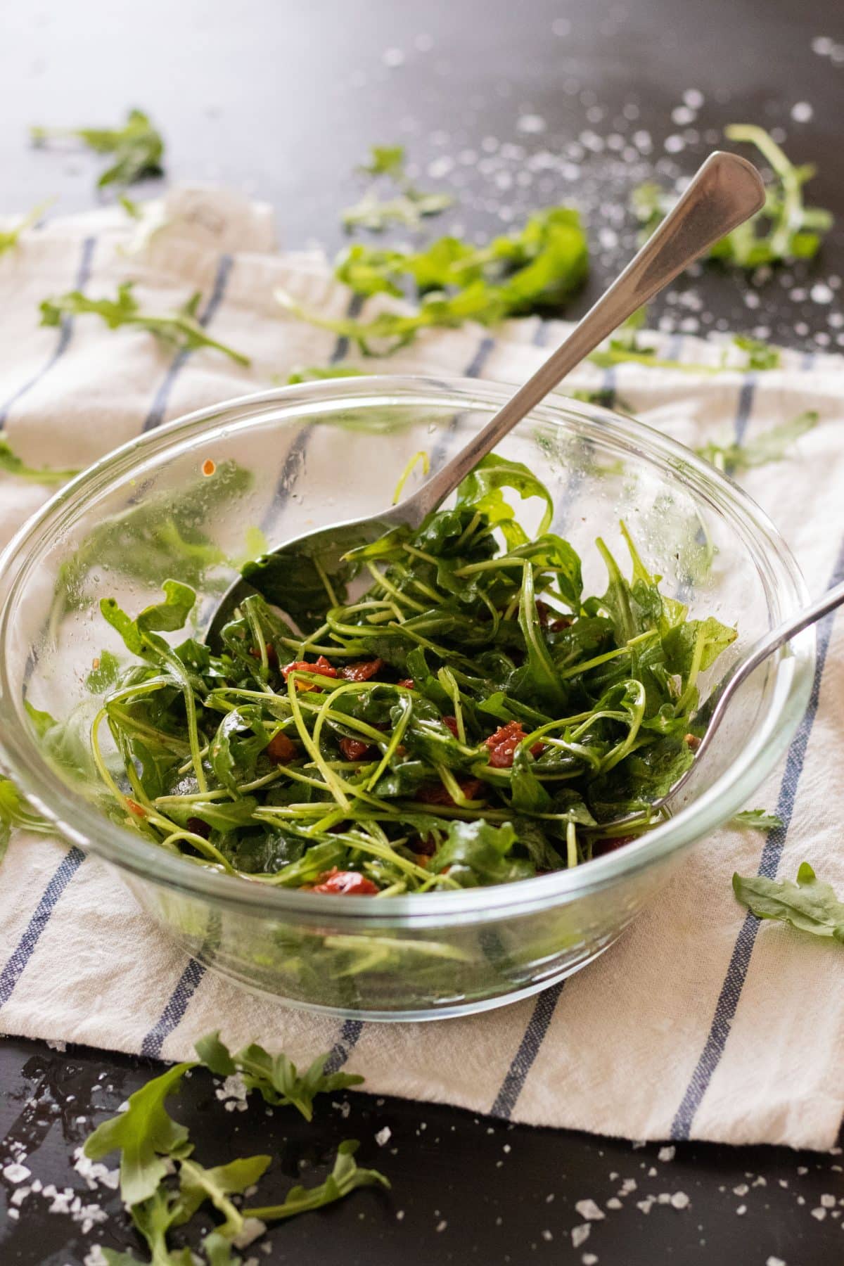 side view of arugula salad with sun-dried tomatoes in a glass bowl with a spoon and fork