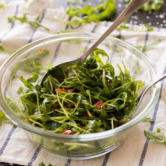 side view of arugula salad with sun-dried tomatoes in a glass bowl with a spon and fork