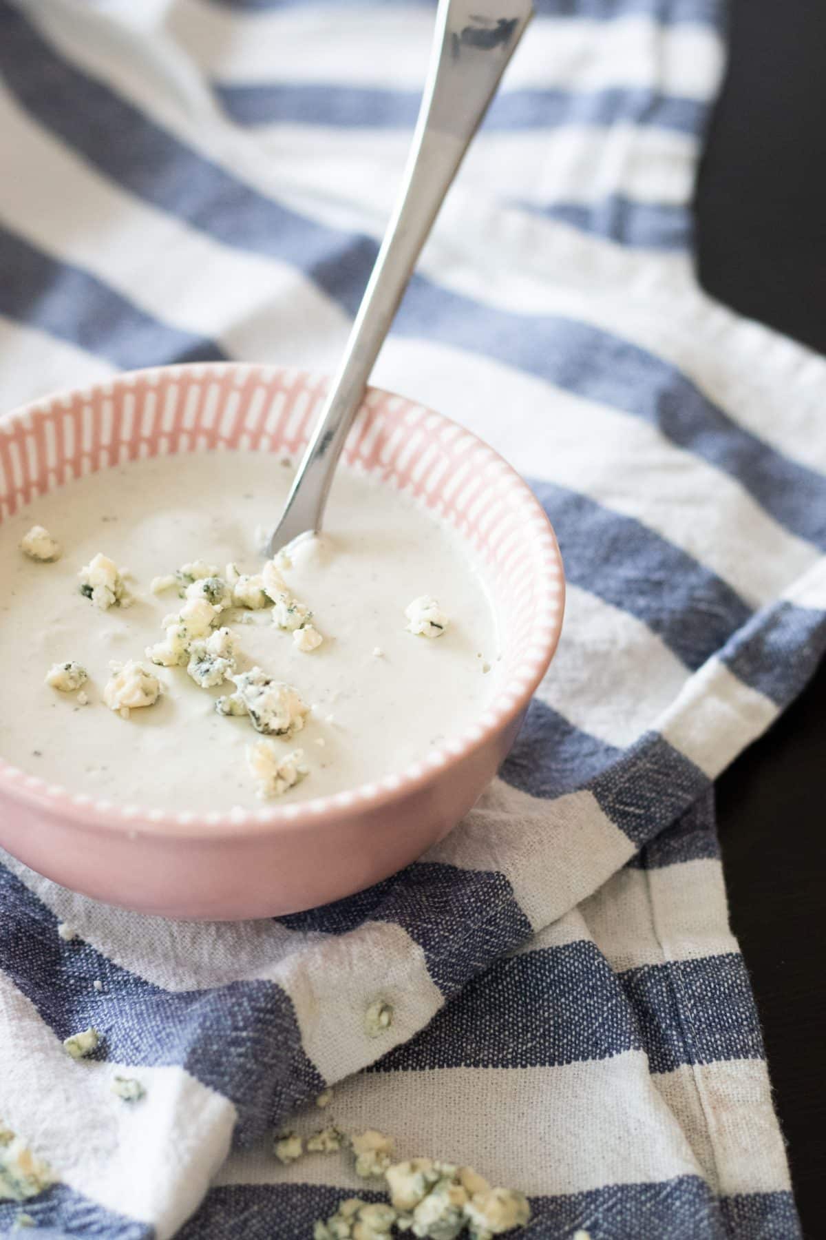 side view of blue cheese sauce and blue cheese crumblesi a pink bowl on a blue and white striped kitchen towel