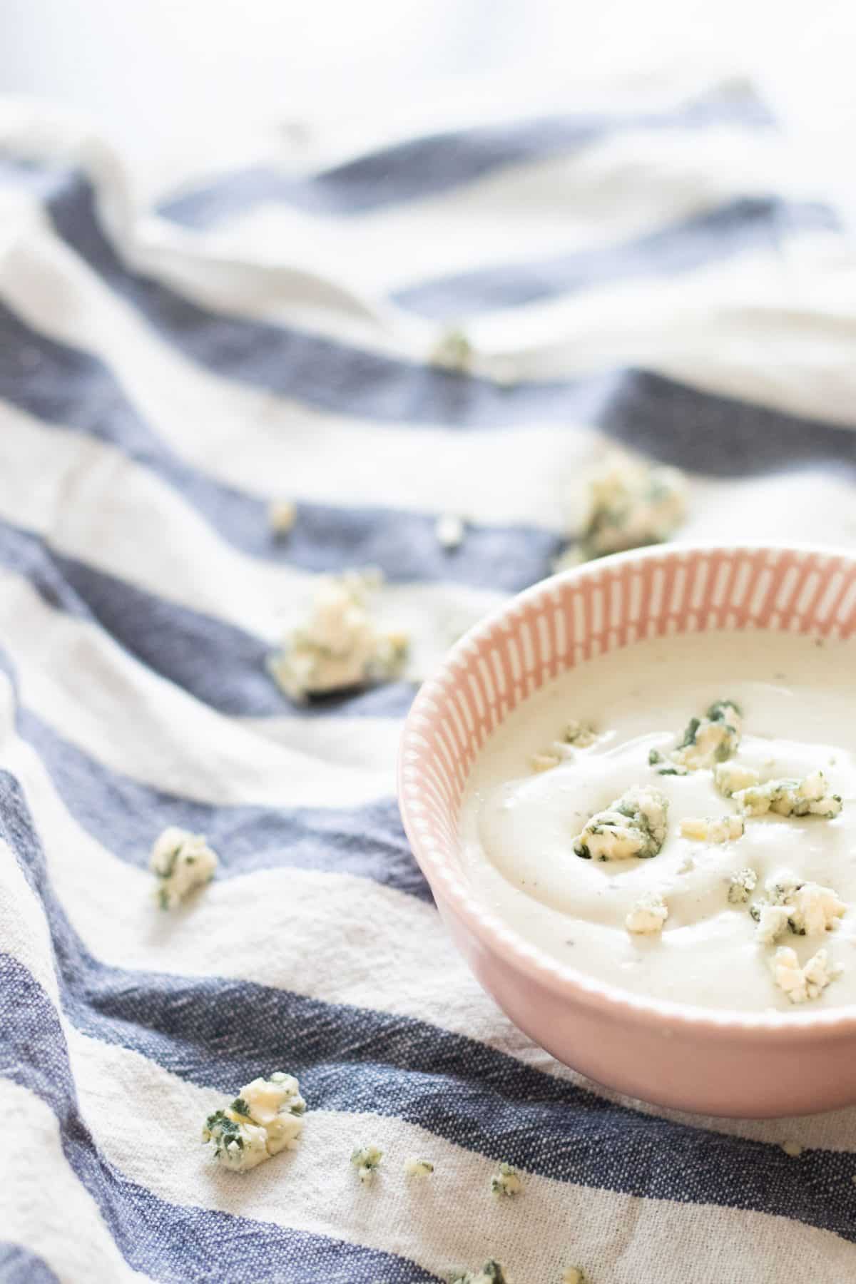 side view of blue cheese sauce and blue cheese crumblesi a pink bowl on a blue and white striped kitchen towel