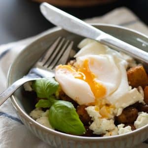 closeup sideview of a bowl with fried potatoes, feta cheese, poached egg and a sprig of basil