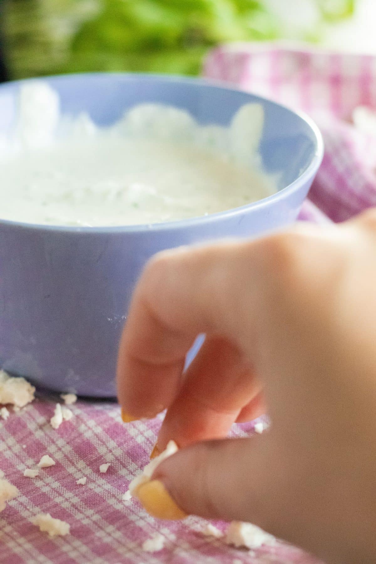 pieces of feta cheese being picked up from a pink and with checkered kitchen towel, in front of a blue bowl with white sauce