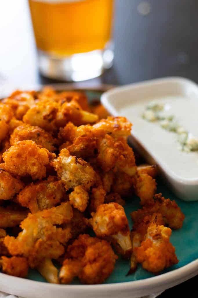 closeup of buffalo cauliflower bites and blue cheese sauce in front of a glass of beer