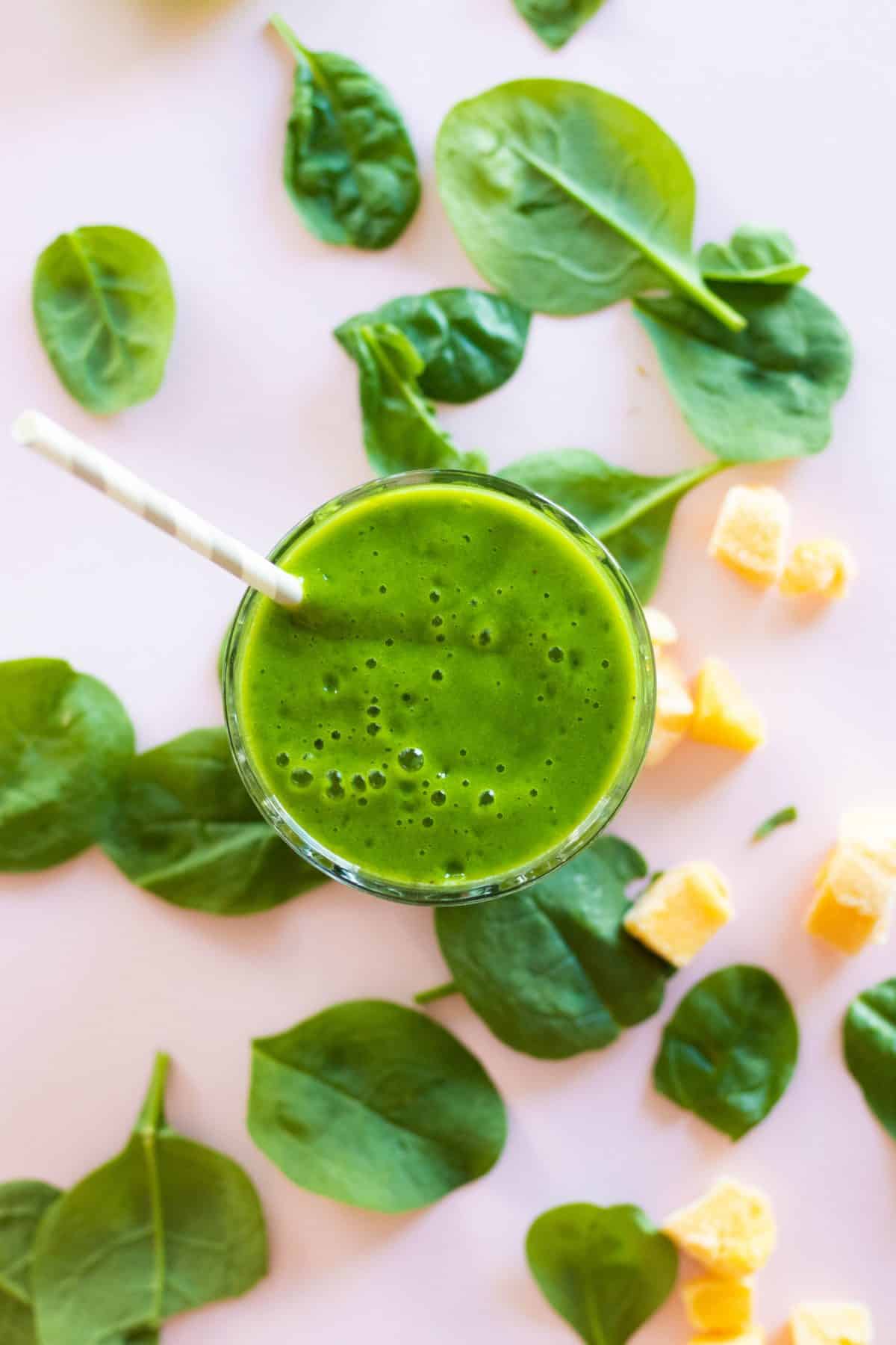 top view of green smoothie in a glass with a straw with mango pieces and spinach leaves around.