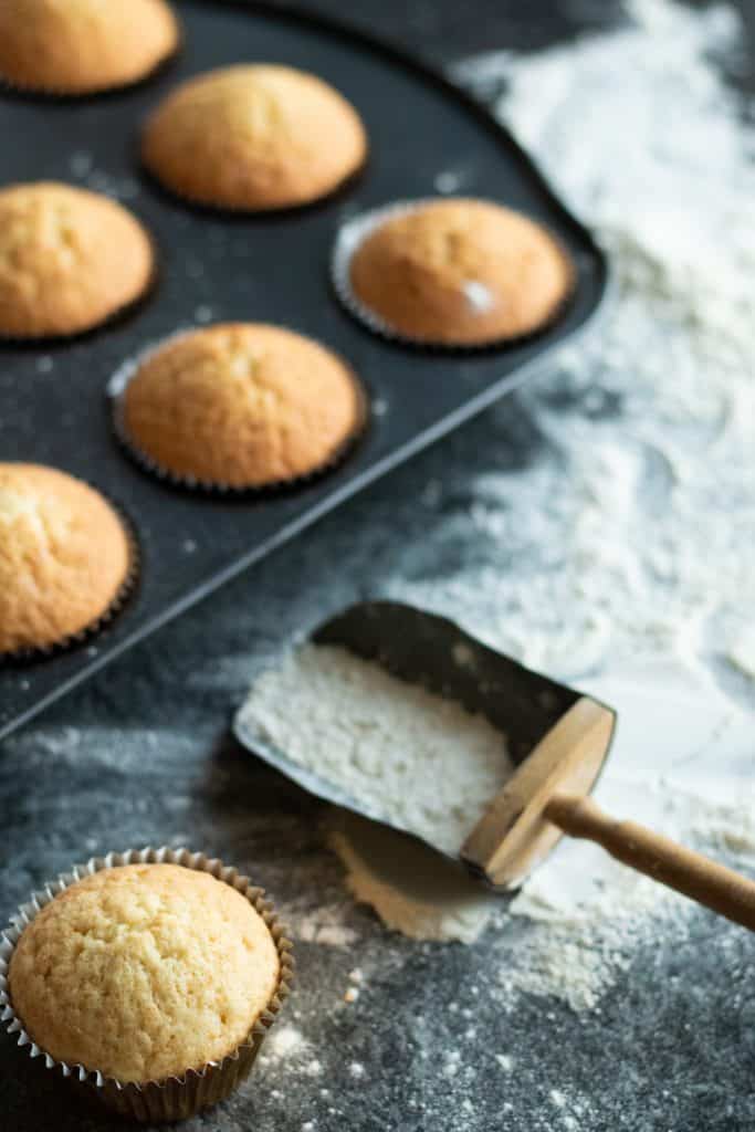 a vanilla muffin in front of a tray of vanilla muffins, with flour on the surface