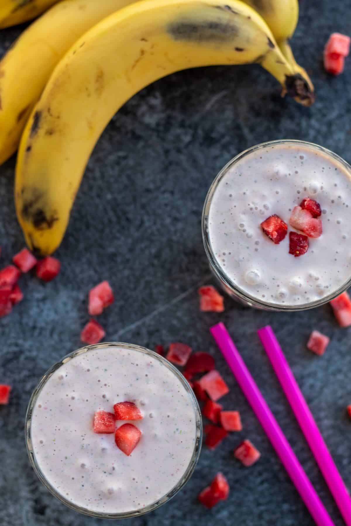 top down view of two strawberry banana smoothies with bananas and frozen starwberries around