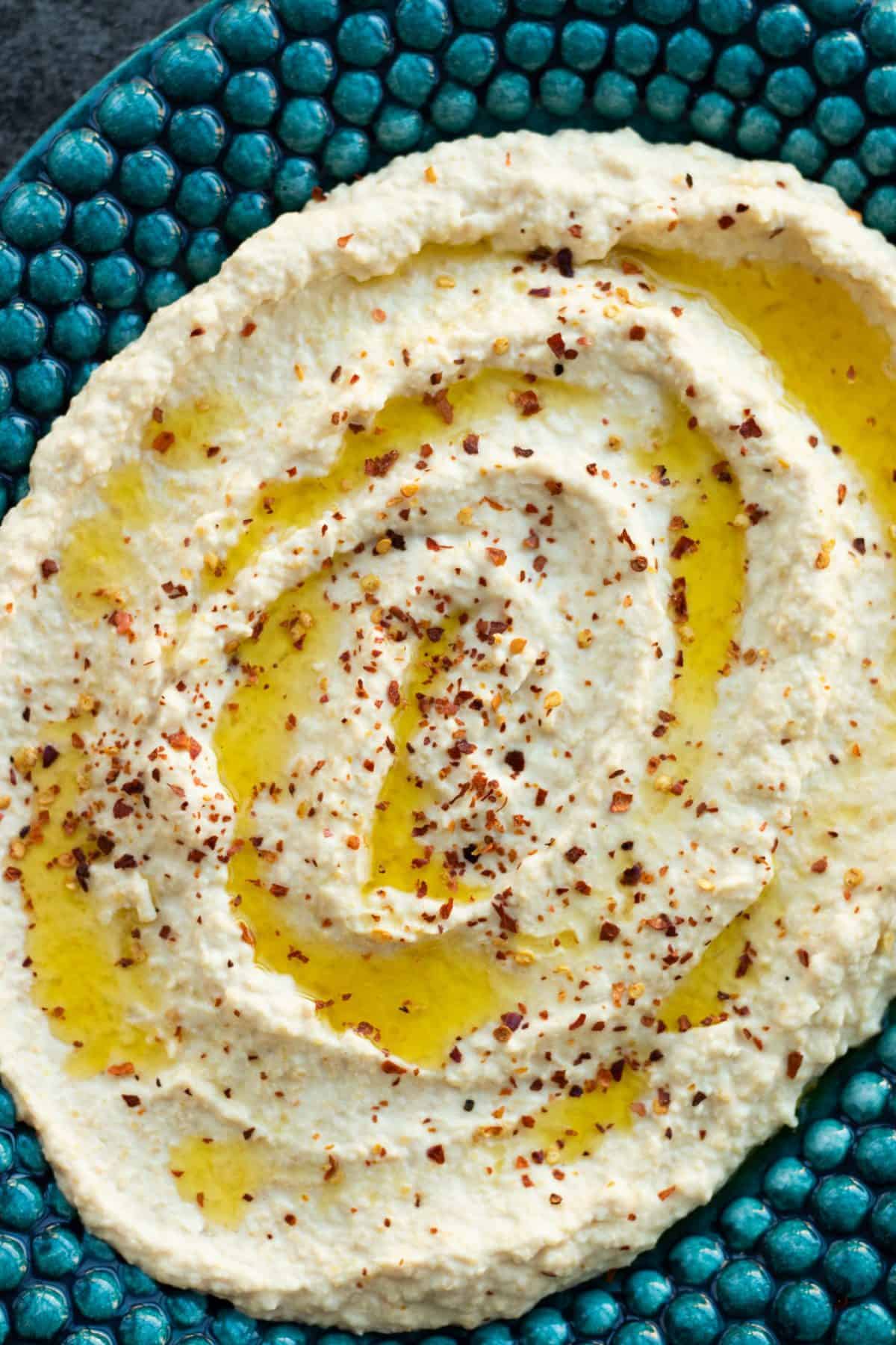 top down view of hummus topped with olive and chili flakes, on top of a blue-green plate