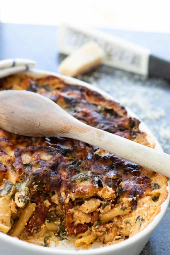 cut through view of pasta bake with spinach and sun-dried tomatoes, with a wooden spoon on top