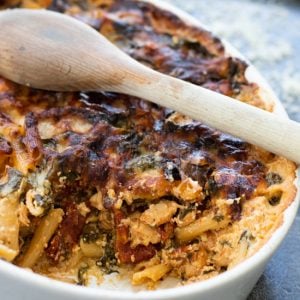 cut through view of pasta bake with spinach and sun-dried tomatoes, with a wooden spoon on top
