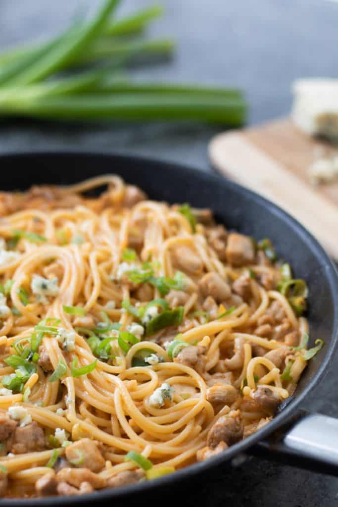 side view of buffalo chicken pasta in a black frying pan with spring oniona and blue cheese in the background