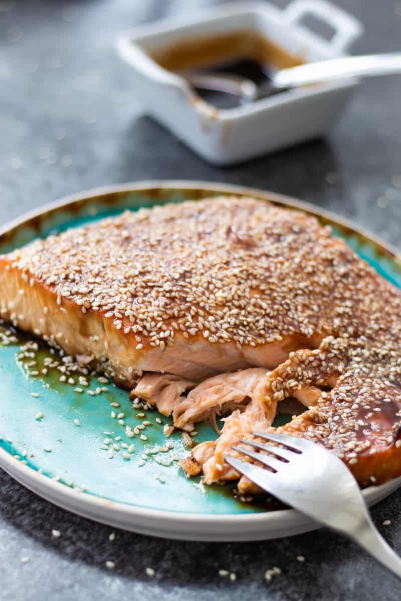 side view of baked teriyaki salmon on a turquoise plate with a fork separating the salmon, and a bowl of teriyaki sauce in the background