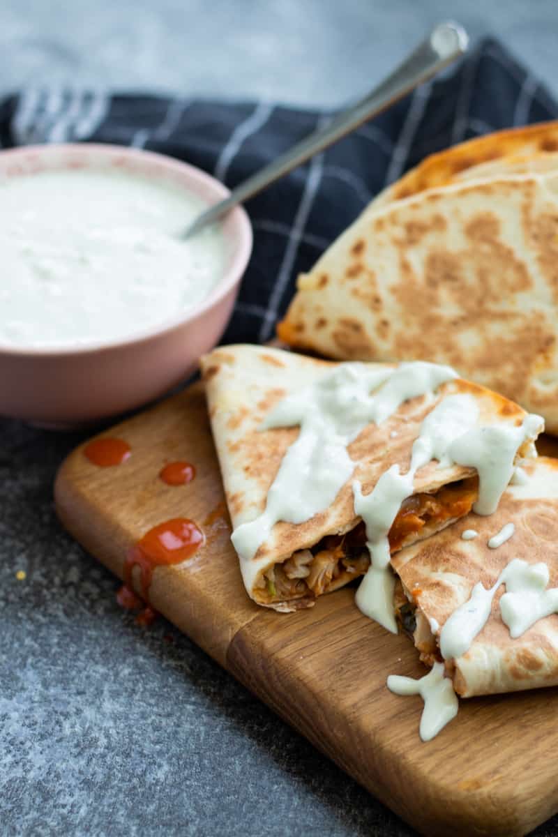 buffalo chicken quesadillas on a wooden chopping board, drizzled with blue cheese sauce and a bowl of blue cheese sauce on the side