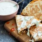buffalo chicken quesadillas on a wooden chopping board, drizzled with blue cheese sauce and a bowl of blue cheese sauce on the side