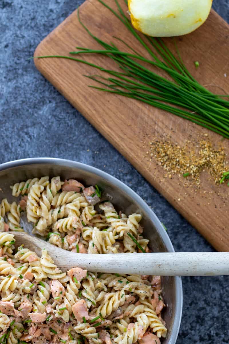 top down image of a pan full of salmon creme fraiche pasta with a wooden spoon, and a chopping board with chives, a lemon and lemon pepper next to it