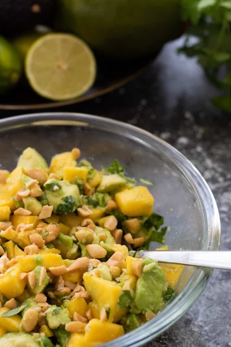side view of a bowl of avocado mango salad with salted roasted peanuts and some sea salt flakes and half a lime in the background