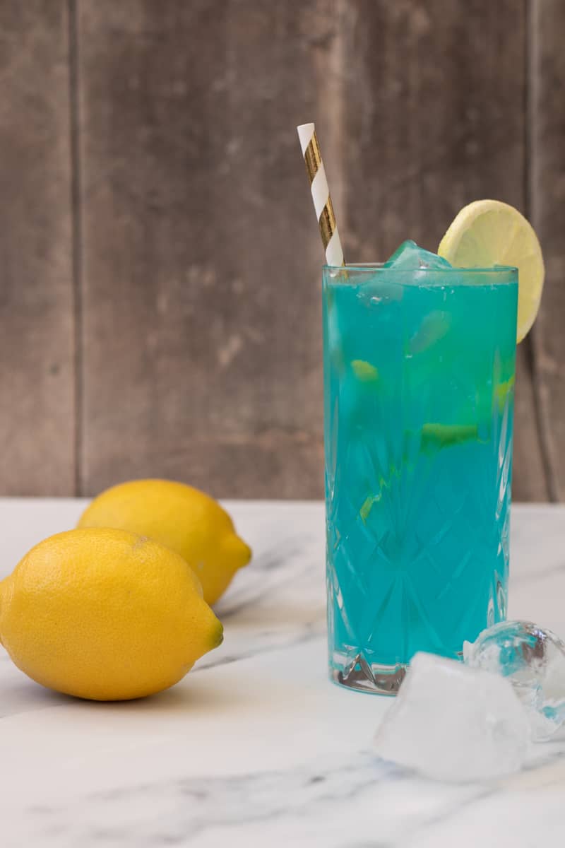 side view of a blue mocktail in a highball glass, decorated with a slice of lemon and a straw, with two lemons and some ice cubes next to it