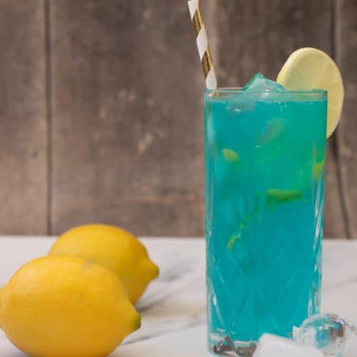 side view of a blue mocktail in a highball glass, decorated with a slice of lemon and a straw, with two lemons and some ice cubes next to it