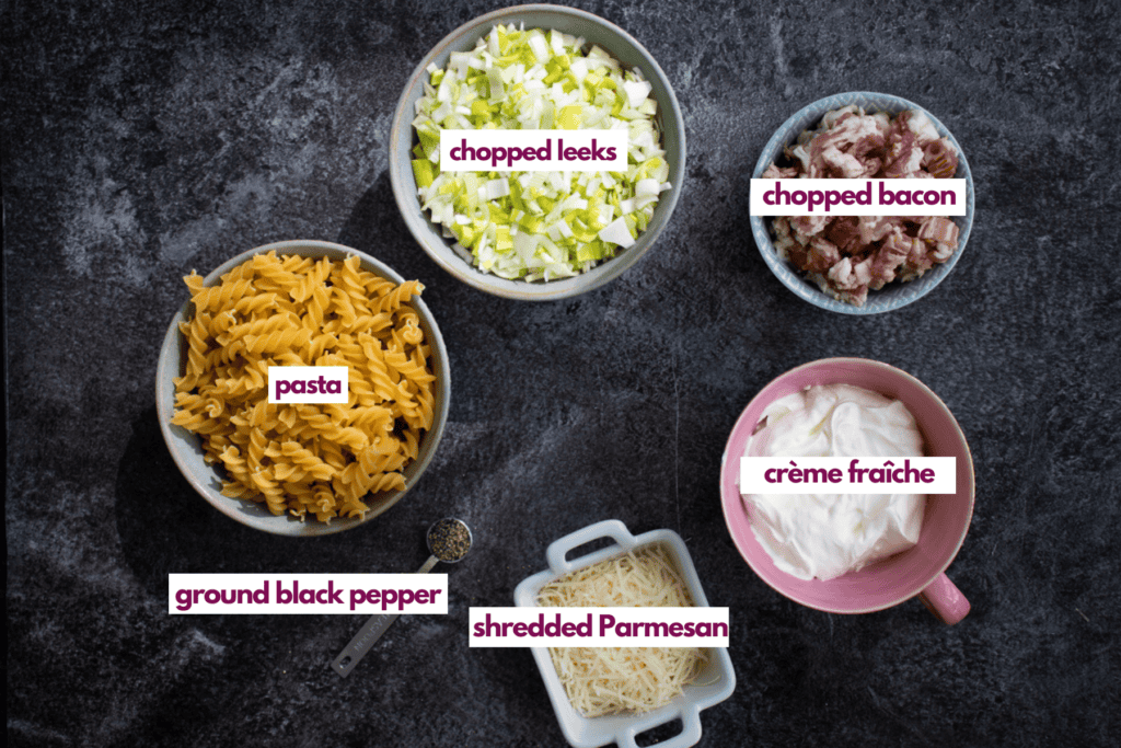 Ingredients needed to make a creme fraiche carbonara including pasta, chopped leeks, chopped bacon, creme fraiche, shredded Parmesan cheese and ground black pepper.