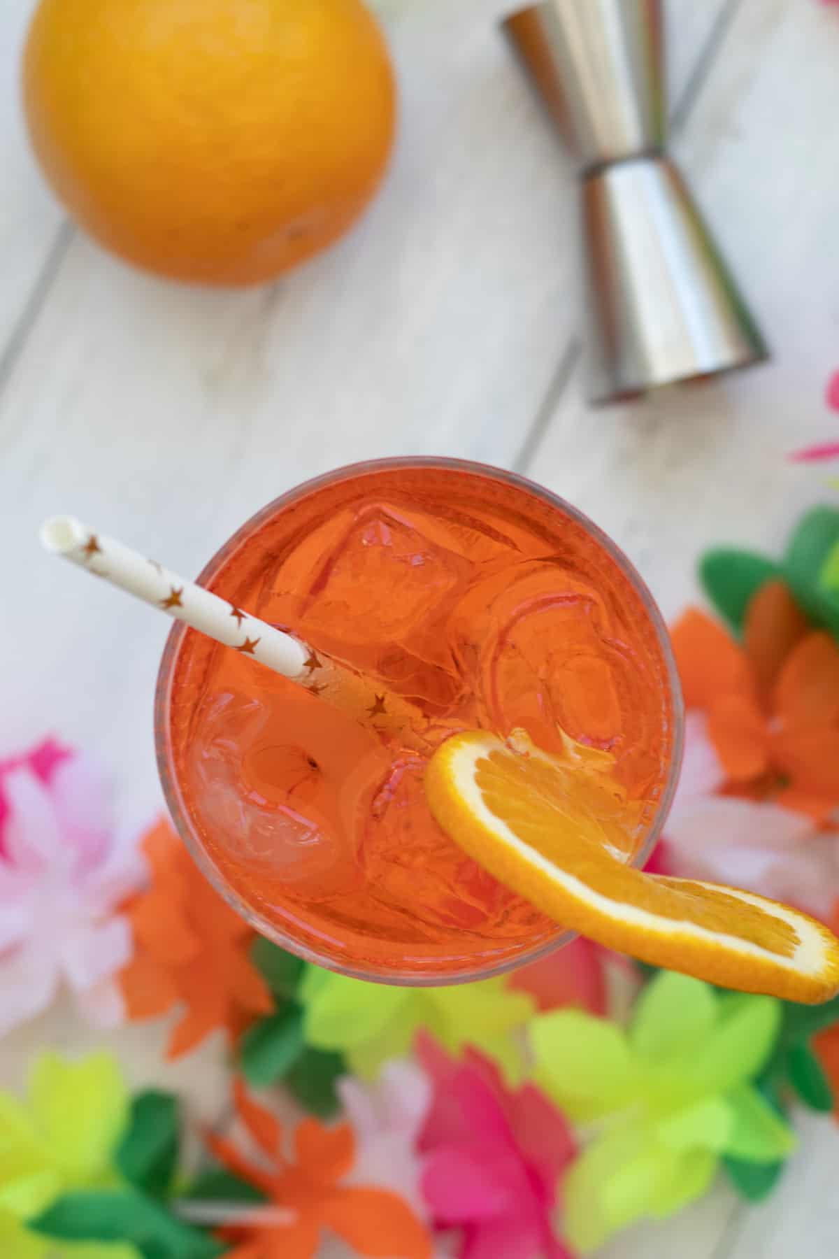 top down view of an aperol spritz garnished with orange and a straw, around it are an orange, a measure and some plastic flowers