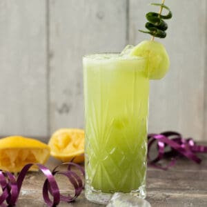 side view of a cucumber tom collins mocktail decorated with cucumber, with lemons in the background