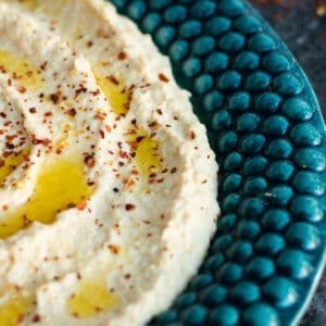 a blue-green plate full of hummus topped with olive oil and chili flakes with a plate of acili ezme in the backgound