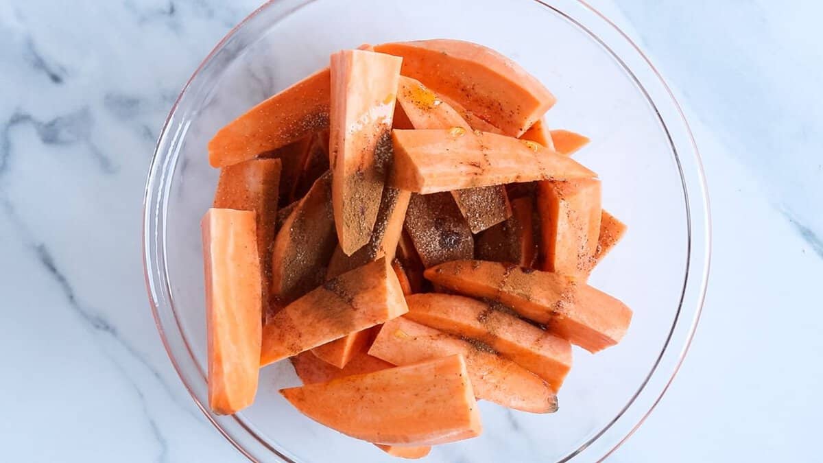 Sweet potato wedges in a glass bowl seasoned with salt, cinnamon and olive oil, before mixing and air frying.