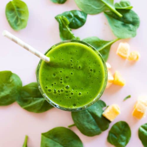 A green smoothie surrounded by mango and spinach.