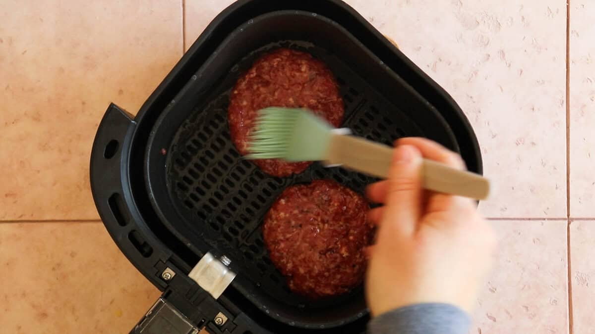 Brushing olive oil on hamburger patties in the air fryer before cooking.