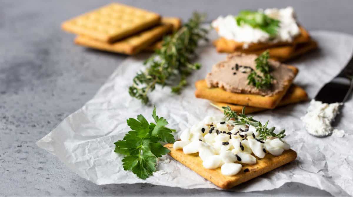 Tasty salted square crackers with cream cheese, cottage cheese, liver pate, seeds and herbs on parchment paper.