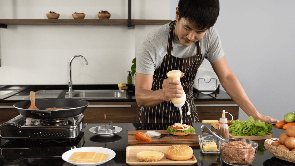 A man squeezing out sauce on a hamburger in a kitchen.