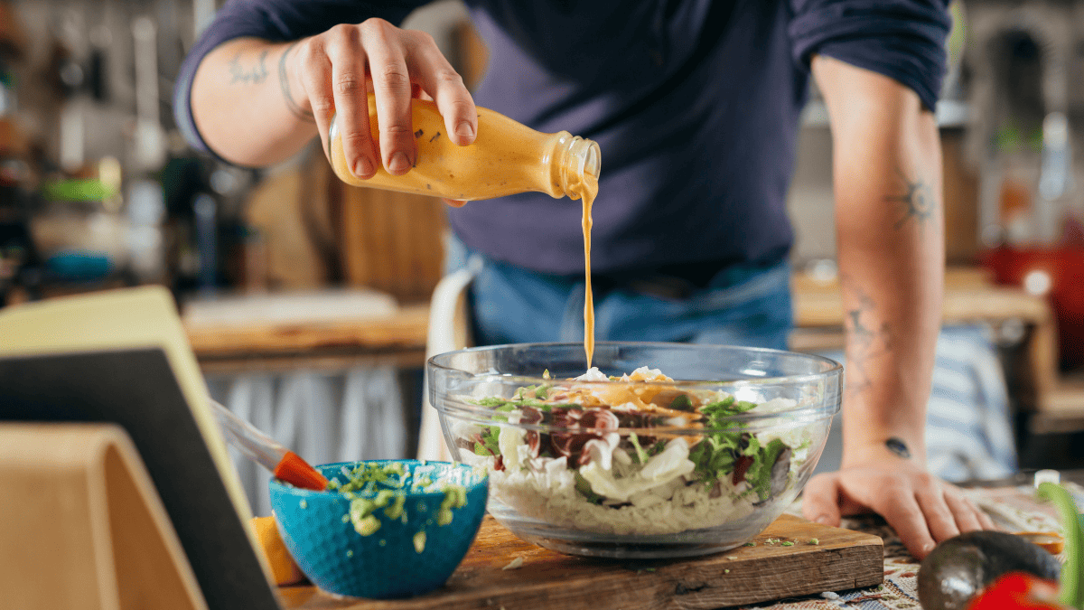 A person pouring salad dressing from a bottle on a salad.